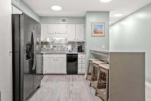 A compact kitchen with white cabinets, a stainless steel refrigerator, a coffee maker, and a wooden stool. - Cozy 2BR condo steps from the beach with ocean views, full kitchen, and great off-season rates (North Myrtle Beach)