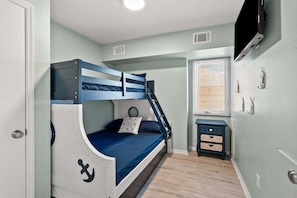 A bunk bed with a nautical theme, a small bedside table, a TV mounted on the wall, and a window with blinds. - Cozy 2BR condo steps from the beach with ocean views, full kitchen, and great off-season rates (North Myrtle Beach)