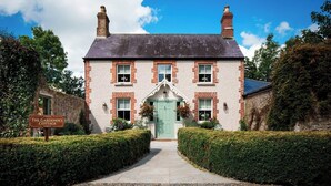 Une charmante maison de campagne traditionnelle en briques, avec une porte verte, une enseigne indiquant « La maison du jardinier » et un chemin de gravier menant à l’entrée.