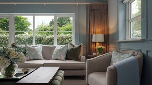 Un salon chaleureux avec un canapé, un fauteuil et une table d’appoint sur laquelle se trouve une lampe. De grandes fenêtres offrent une vue sur la verdure environnante.