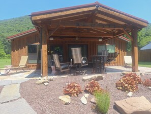 Une cabane en bois dotée d’un patio couvert, aménagé avec des chaises et une table, et entourée d’un espace recouvert de gravier, de rochers et de plantes.