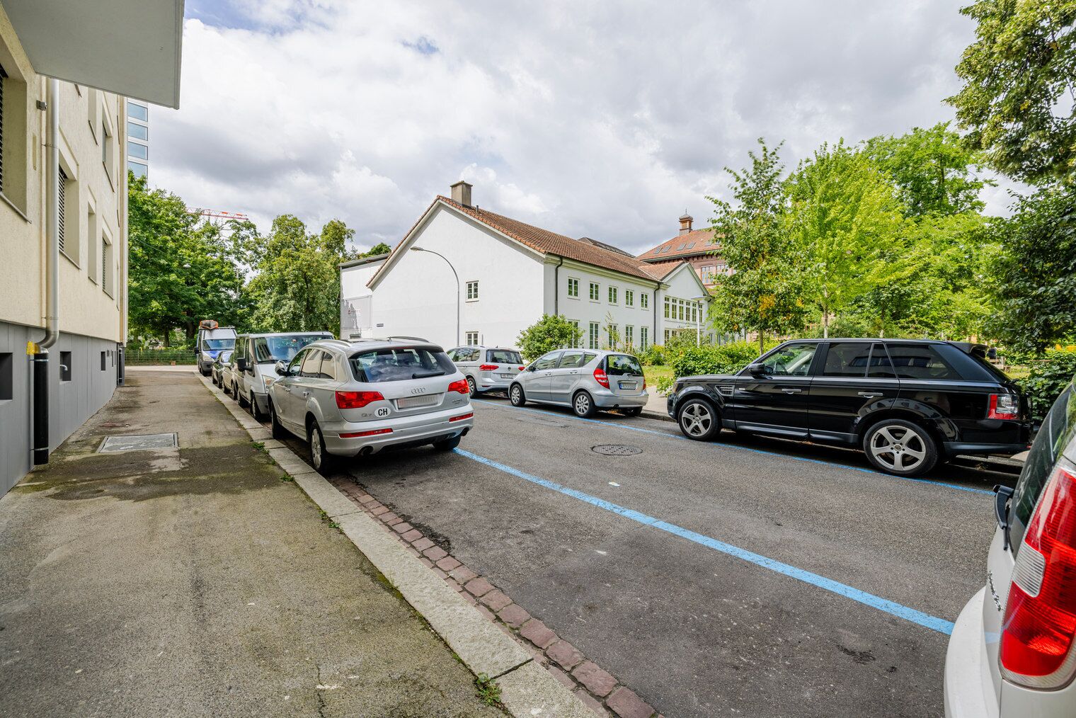 A residential street with parked cars, a white house, and a red-roofed building.
