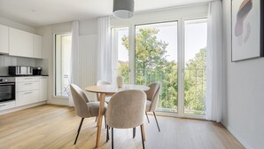 A modern kitchen with white cabinets, a microwave, and a coffee maker. There is a dining area with a round table and four chairs. Large windows provide a view of trees and buildings outside. - Blueground | Basel-Vorstaedte, view, nr Rhine (Basel)