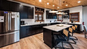 A modern kitchen with dark cabinets, stainless steel appliances, a wooden ceiling, and pendant lighting. - Luxury Mountain Lodge | Huge Game Room | Htd Pool (Sevierville)