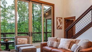 A cozy living room with a leather sofa, a coffee table, and a view of a lush garden through large sliding glass doors. - Luxury Mountain Lodge | Huge Game Room | Htd Pool (Sevierville)