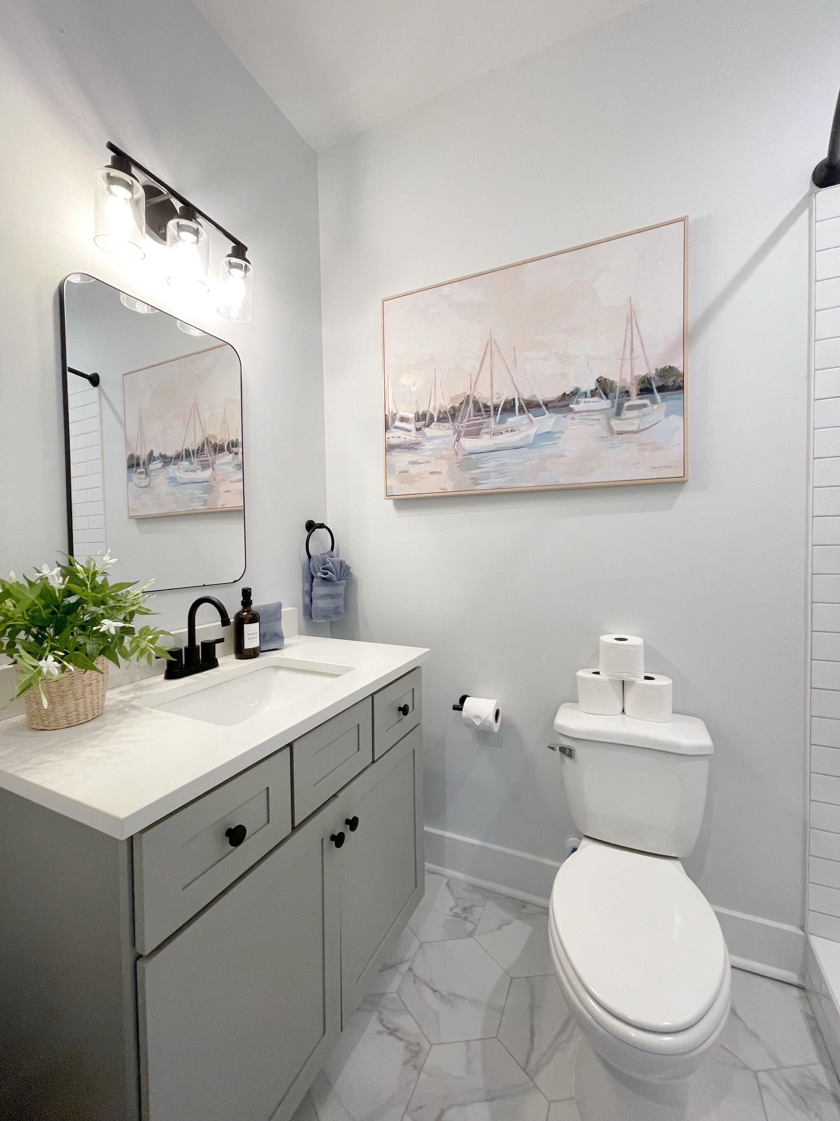 A bathroom with a white sink, a mirror, a toilet, and a painting of sailboats on the wall.