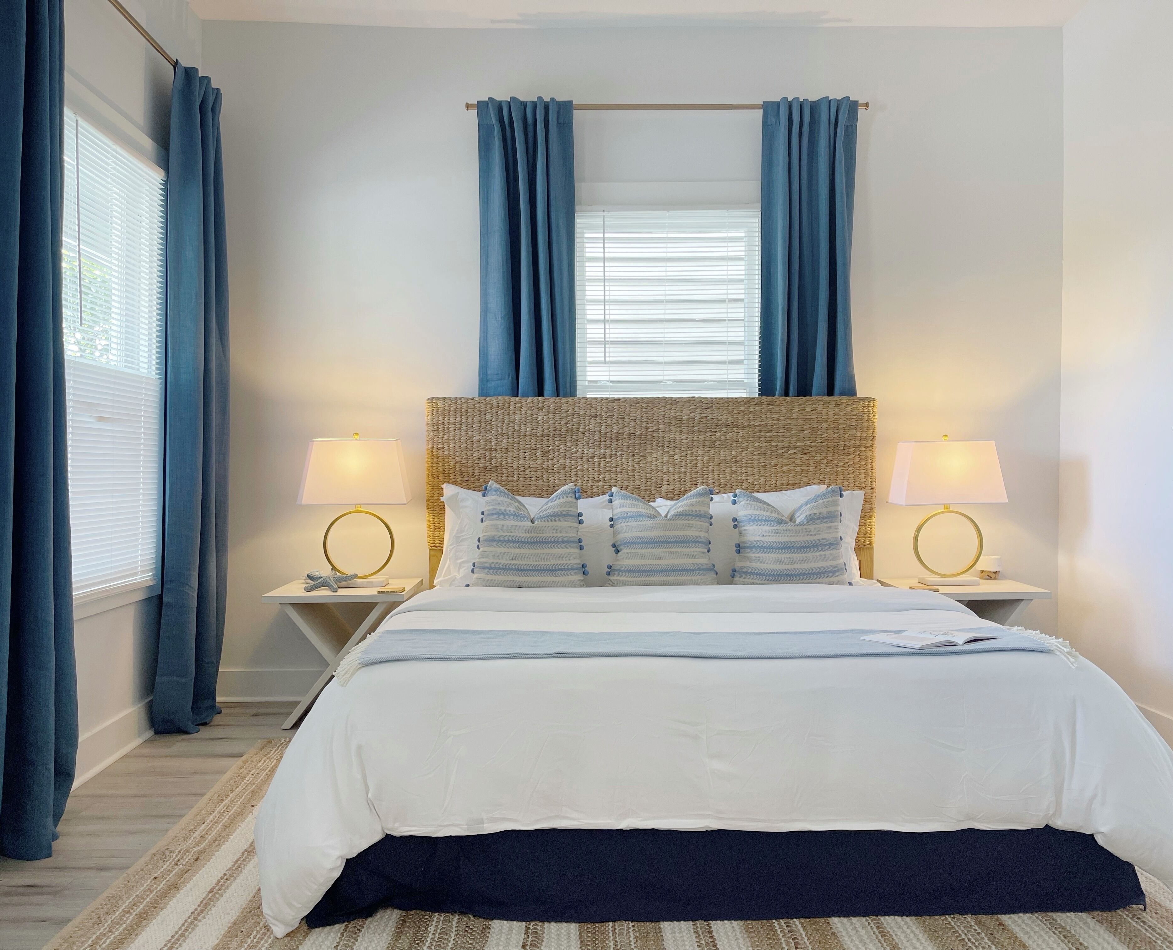 A neatly arranged bedroom with a large bed, two bedside tables with lamps, a wicker headboard, and blue curtains.