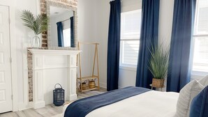 A bedroom with a white fireplace, a bed with a blue bedspread, and a large window with blue curtains. - Stylish 1900s Renovated Apartments (Savannah)