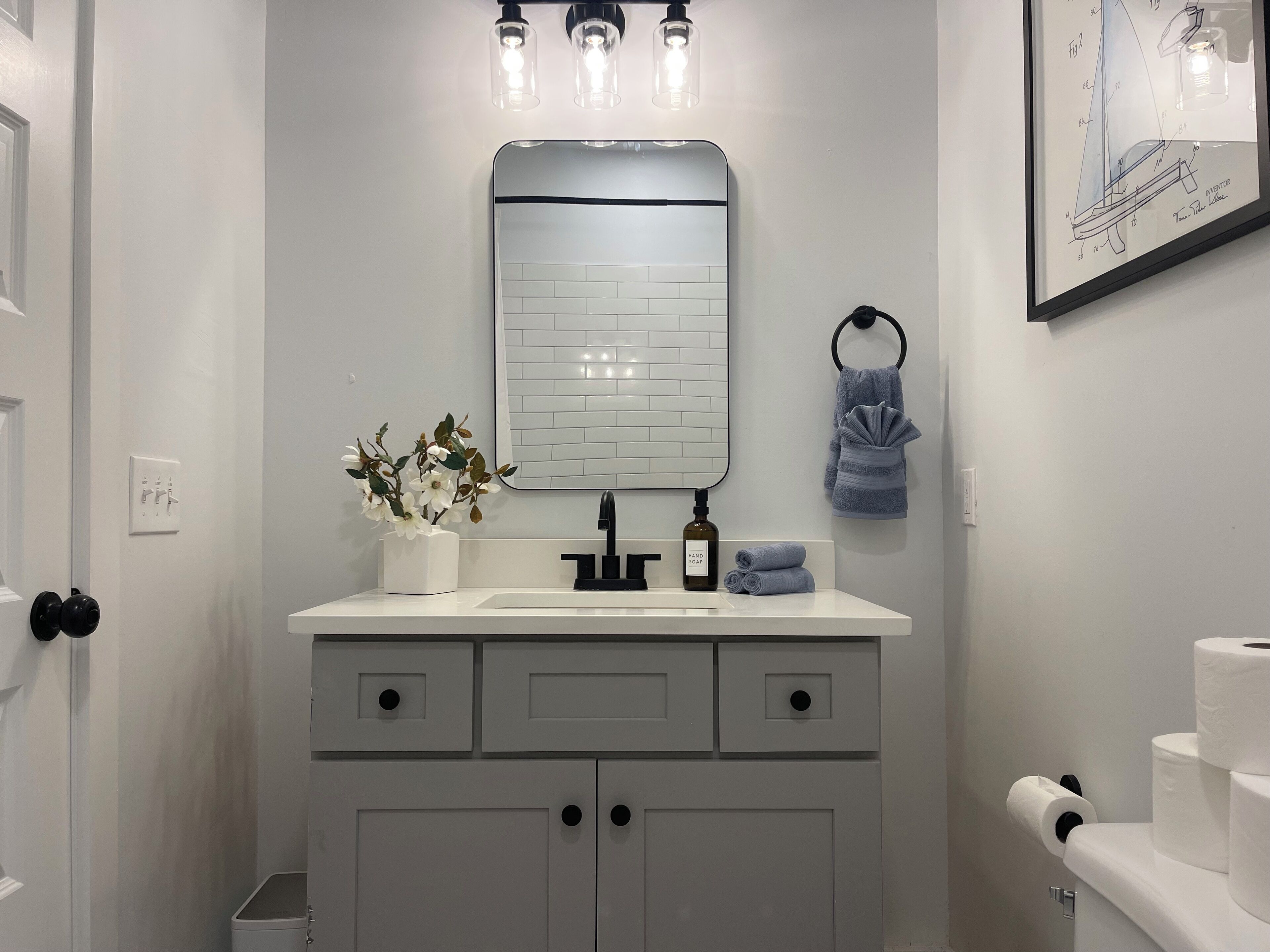 Une salle de bain avec un meuble-lavabo blanc, un miroir, un porte-serviettes, une plante et un dessin de brevet encadré.