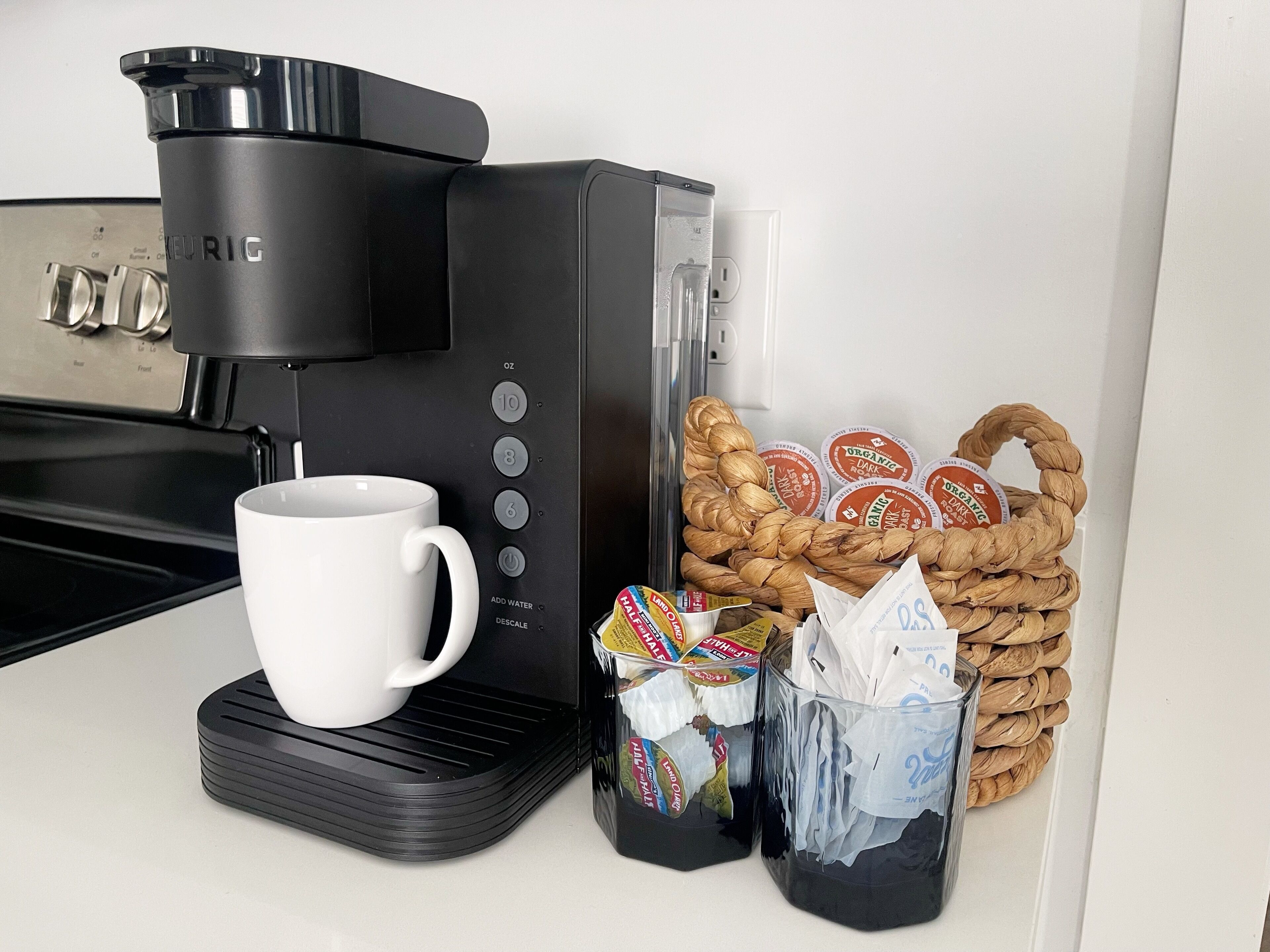 A coffee maker, a white mug, a basket of snacks, and a container with a straw.