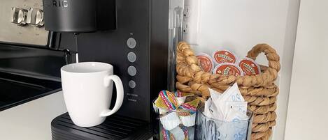 A coffee maker, a white mug, a basket of snacks, and a container with a straw.