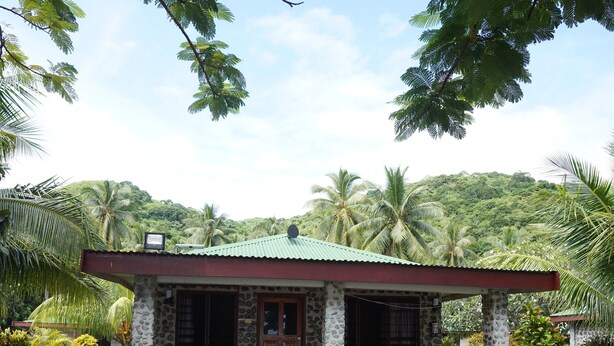 A small stone building with a red roof and a porch, surrounded by greenery and palm trees. - Korovou Eco Resort (Naviti Island)