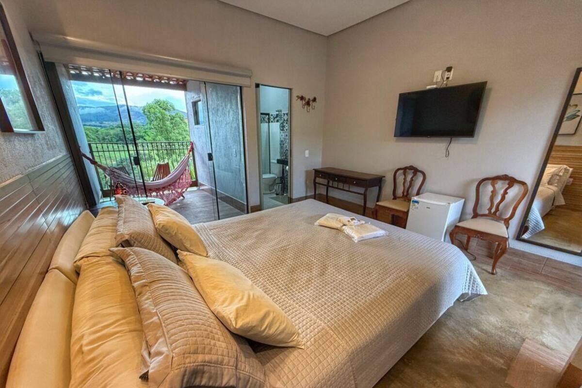 A bedroom with a large bed, a desk, a chair, a TV, and a balcony with a hammock.