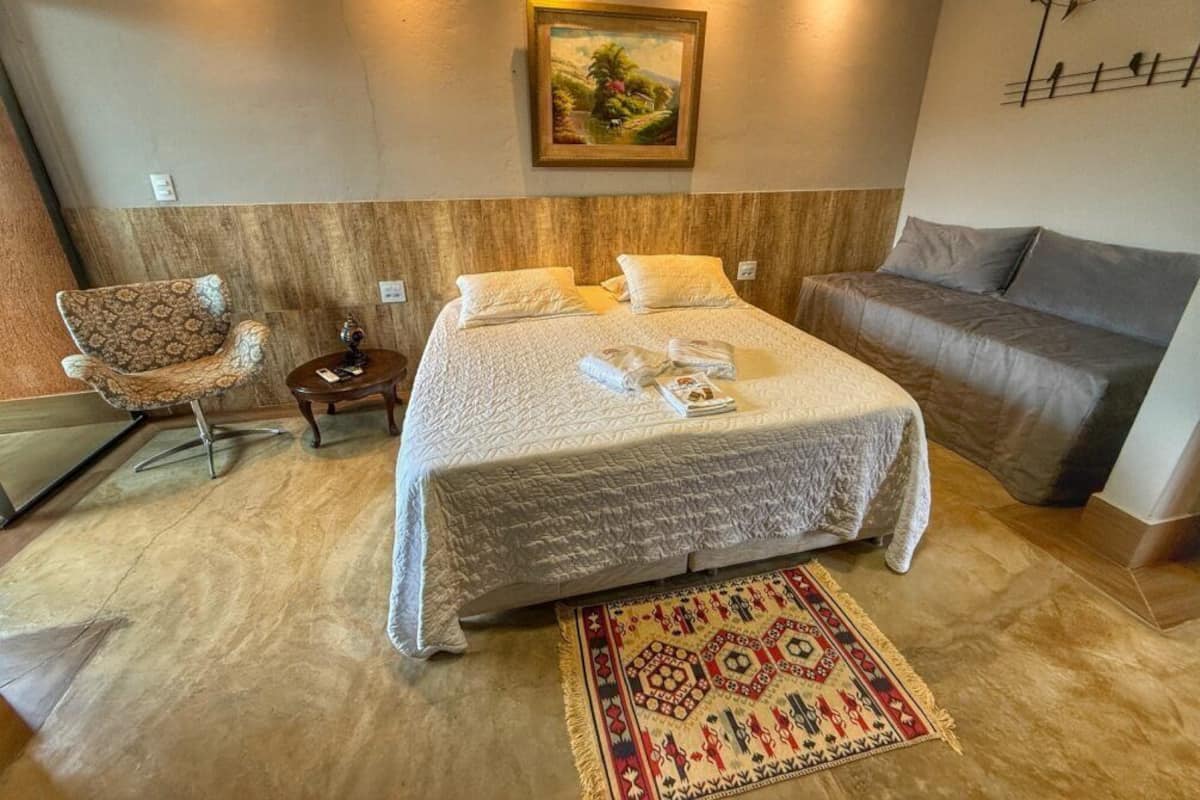 A bedroom with a large bed, a sofa, a chair, a nightstand, and a painting on the wall.