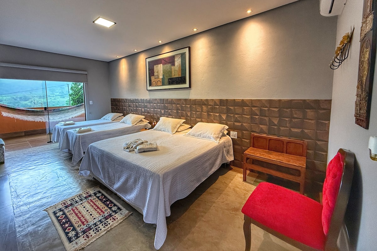 A hotel room with three beds, a red chair, a wooden bench, and a large window offering a view of the outdoors.