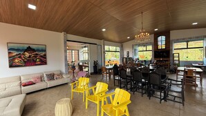Un vaste espace de vie doté d’un plafond en bois, d’un grand tableau, d’un canapé, de fauteuils jaunes et d’un coin repas avec des tables et des chaises.