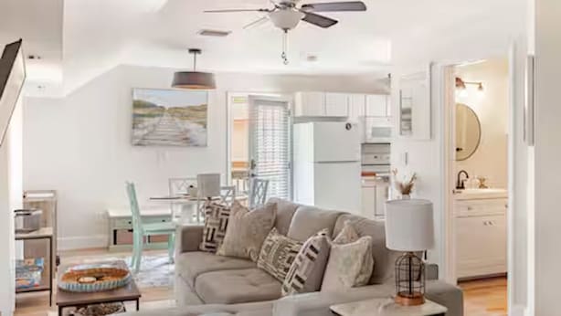 Una sala de estar moderna con un sofá gris, un comedor con mesa y sillas, y una cocina con armarios blancos.