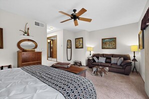 A bedroom with a bed, a wooden dresser, a ceiling fan, a brown leather sofa, two wall-mounted lamps, and a door leading to another room.