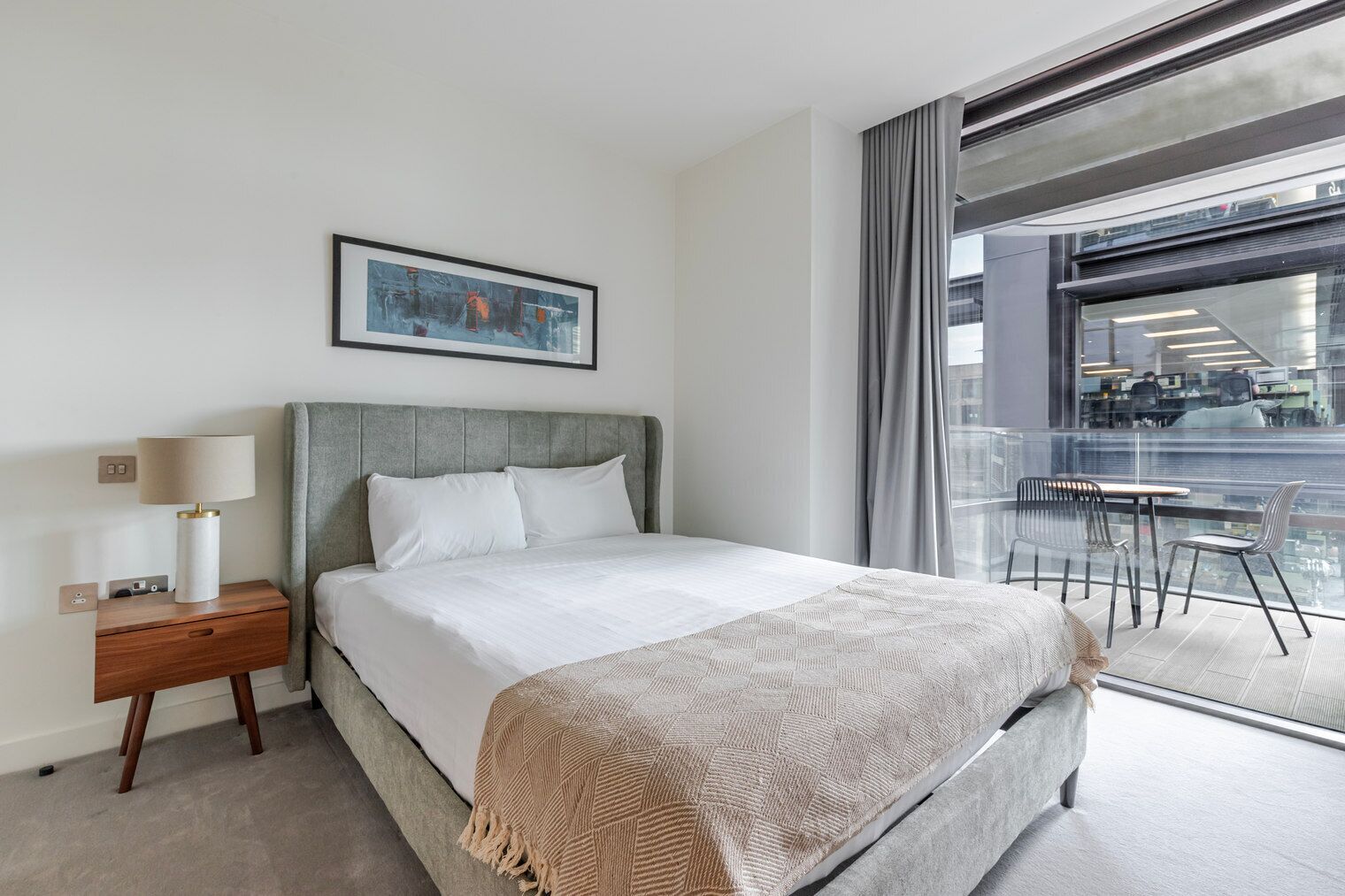 Une chambre d’hôtel moderne avec un grand lit, une table de chevet avec une lampe, un tableau abstrait encadré au mur et une vue sur la ville par une grande fenêtre.
