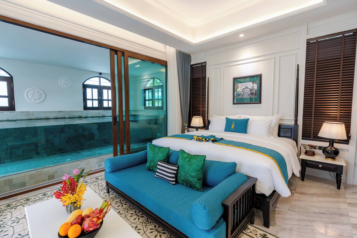 Une chambre d’hôtel avec un grand lit, un canapé, une petite table avec des fruits et une vue sur une piscine.