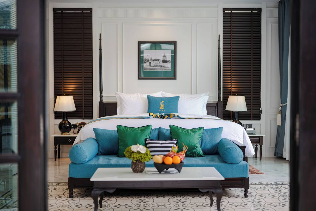 Une chambre d’hôtel avec un grand lit, deux tables de chevet, une table basse avec des fruits et des fleurs, et un tableau encadré au mur.