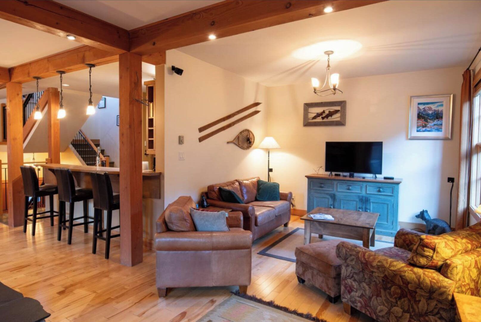 Una sala de estar acogedora con un sofá, un sillón estampado, una mesita de centro y un televisor. Una escalera conduce a otro nivel, y un comedor con sillas y una mesa.