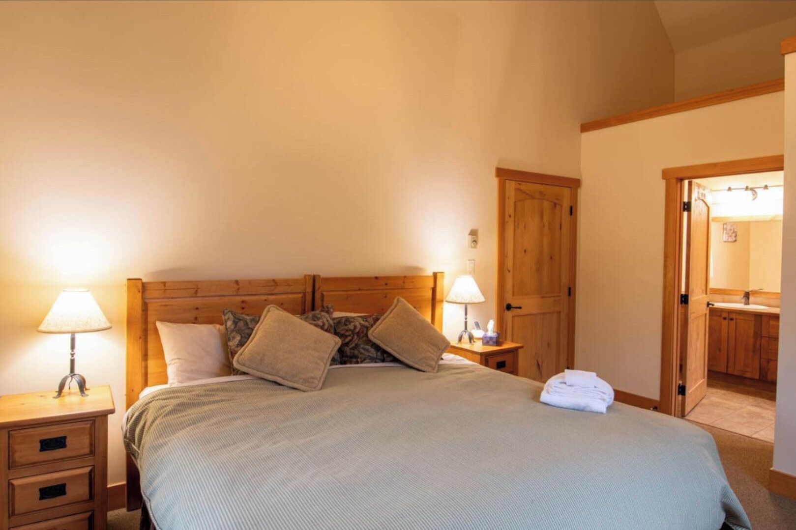 Une chambre à coucher avec une tête de lit en bois, deux tables de chevet avec des lampes, un lit recouvert d’une couverture verte, et une salle de bain visible à travers une porte ouverte.
