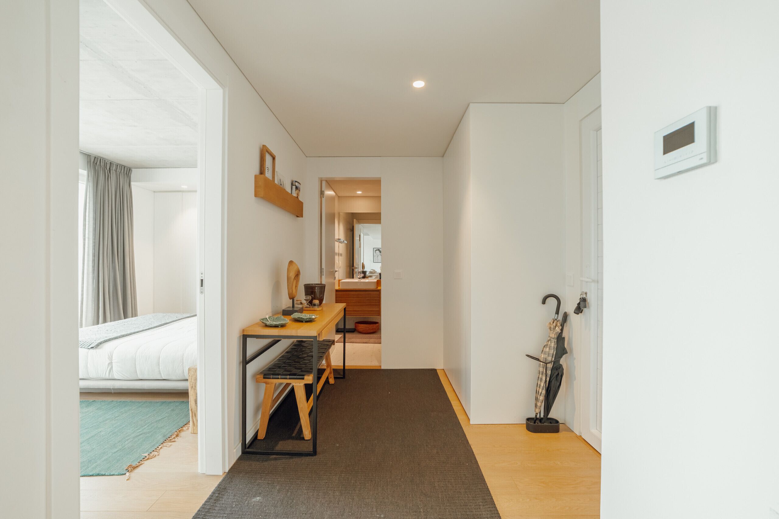 A hotel room with a bed, a desk, a chair, a small table, a mirror, a wall-mounted shelf, and a door leading to a bathroom.