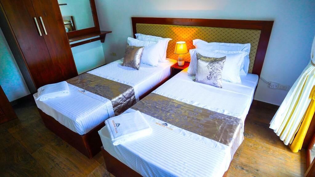 A hotel room with two single beds, a nightstand with a lamp, wooden wardrobe, and a mirror.