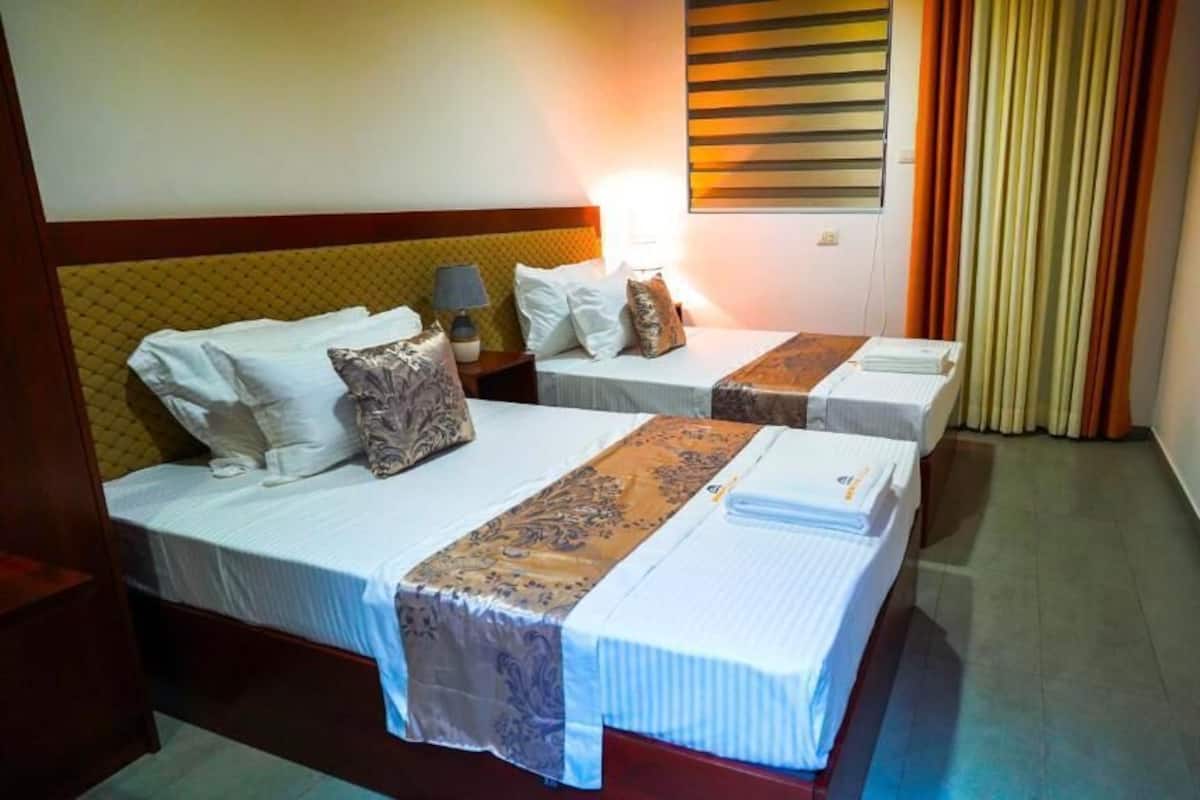 Ein Hotelzimmer mit zwei Betten, jeweils mit Kopfteil, Nachttischen, Lampen und Vorhängen.