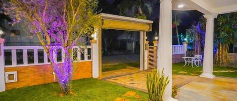 A covered outdoor area with a white column structure, a small garden with grass and potted plants, and a patio area with a table and chairs.