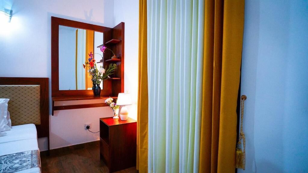 A hotel room with a bed, a bedside table with a lamp, a mirror, and a window with yellow curtains.