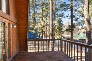 Una terraza de madera con barandilla metálica, con vista a una zona boscosa con árboles altos y un parque infantil.
