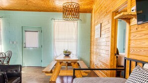 A cozy cabin-style room with a wooden ceiling, a checkered sofa, a wooden dining table with a bowl of fruit, and a window with blinds. - Crystal Clear Frio River - Private Waterfront House - Room for the Family (Concan)