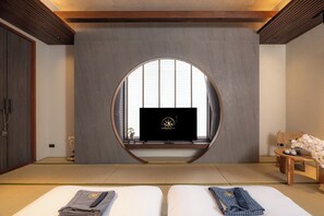 Una habitación con suelo de tatami, un televisor sobre una estantería, un banco de madera y una cama con ropa doblada cuidadosamente.