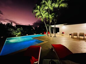 Une piscine avec des chaises longues et un espace salon couvert, éclairé le soir.