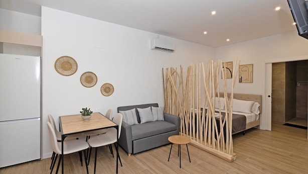 A modern, well-lit room with a grey sofa, a wooden dining table, and a small coffee table. There is a bed with a wooden headboard and a white wall with three woven wall hangings.