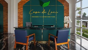 A reception desk with a green backdrop and a logo, two blue chairs, and a view of greenery through large windows. - Casa De Lani Resort & Convention (Baturaden)