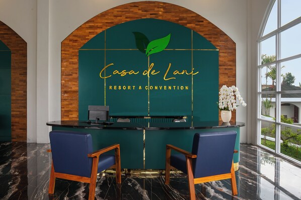 Un mostrador de recepción con fondo verde y un logotipo, dos sillas azules y vistas a la vegetación a través de grandes ventanales.