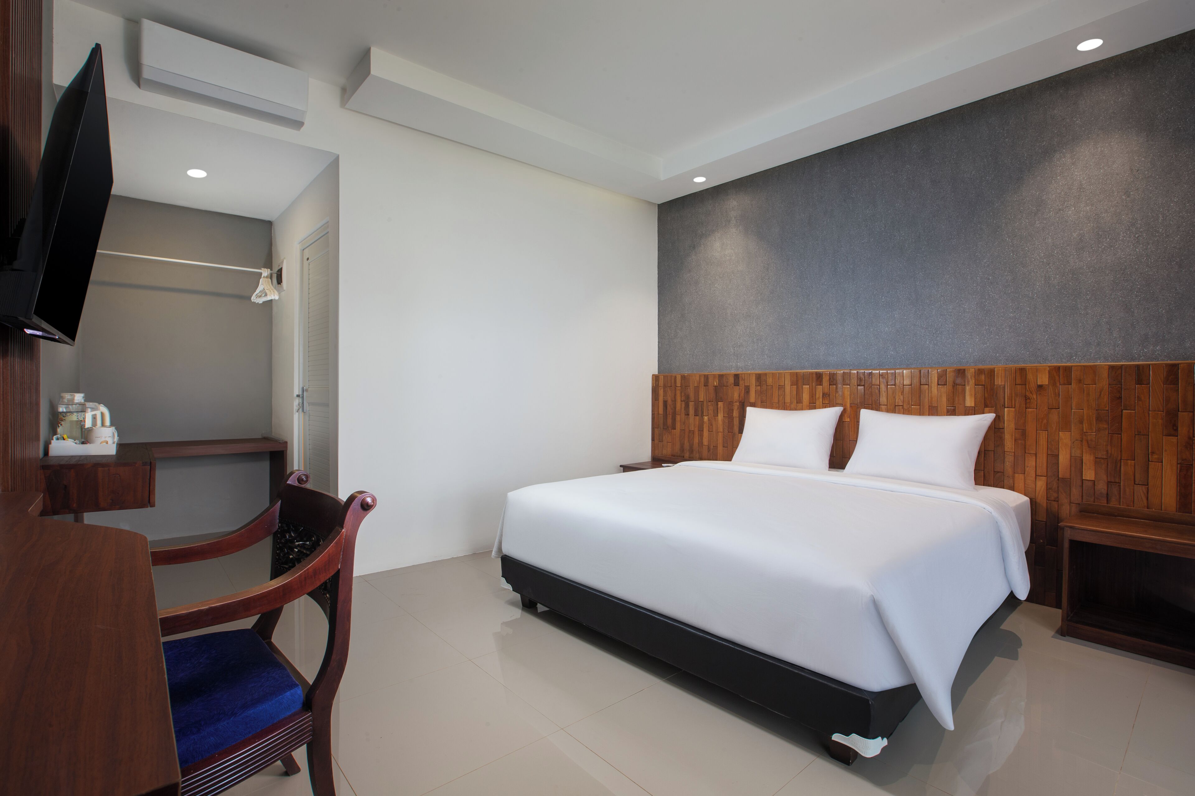 A modern hotel room with a large bed, wooden headboard, a flat-screen TV, a wooden chair, and a bedside table.
