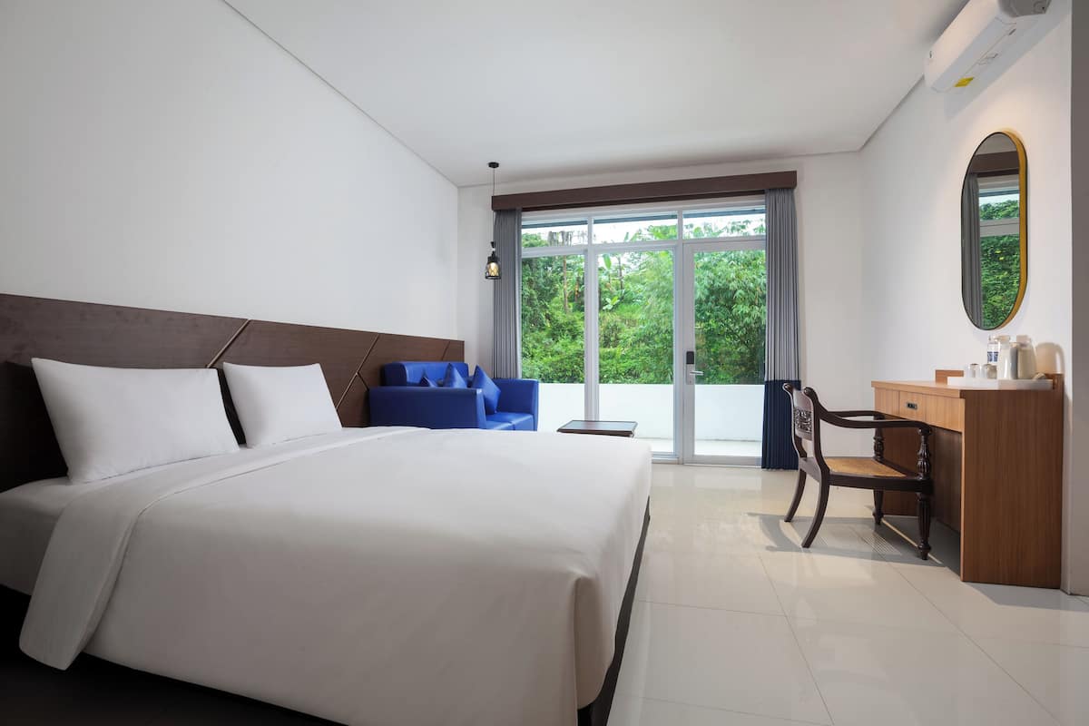 Habitación de hotel con una cama grande, un sofá azul, un escritorio de madera y vistas a la vegetación por la ventana.