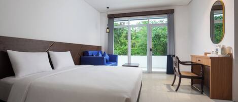 A hotel room with a large bed, a blue sofa, a wooden desk, and a view of greenery through the window.
