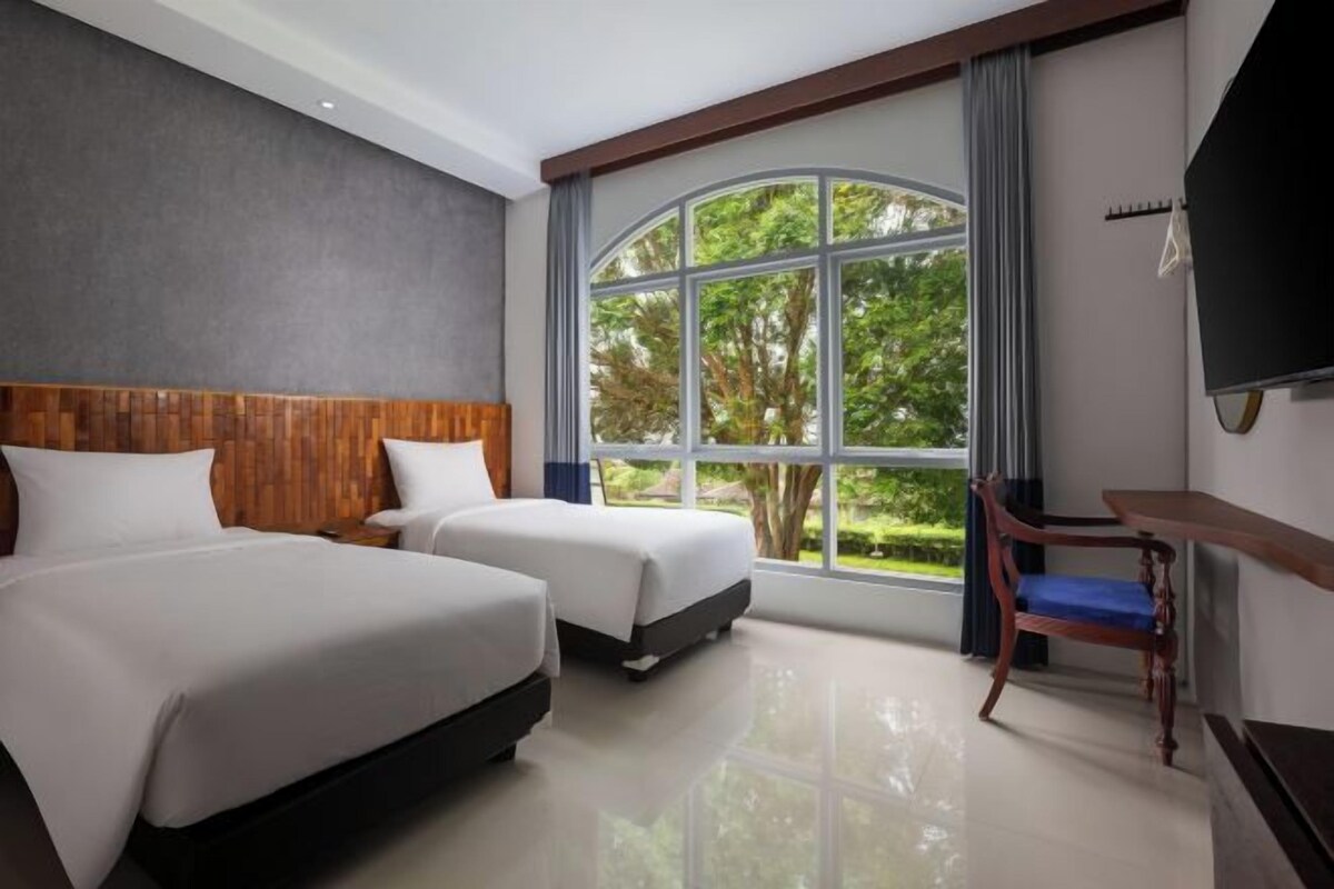 Une chambre d’hôtel avec deux lits, une tête de lit en bois, une grande fenêtre donnant sur un espace verdoyant, une télévision et un bureau avec une chaise.