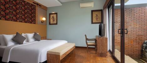 Quarto de hotel com cama, um banco de madeira, uma cadeira e uma grande porta de vidro deslizante que dá para uma área externa.