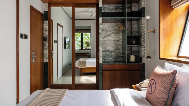 A bedroom with a bed, a wooden wardrobe, a mirror, a nightstand, and a window with curtains. - The Kin Hotel (Mumbai)