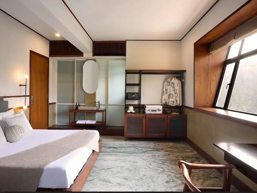 Habitación de hotel con cama, tocador con espejo, cómoda, ventana con vistas y un pequeño rincón de descanso.