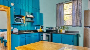 A kitchen with blue cabinets, a wooden table, and a window with checkered curtains. - Beautiful Secluded Cabin in the Woods - Second Wind Ranch (Leakey)
