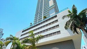 A modern high-rise building with multiple balconies, surrounded by palm trees and greenery.