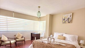 A spacious bedroom with a large bed, two chairs, a desk, and a window with blinds.