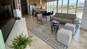 Ein modernes Wohnzimmer mit Sofa, Hocker und Couchtisch, ein Essbereich mit Stühlen und eine Küche mit Geräten.